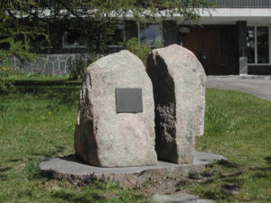 Kahteen osaan haljennut kivi ison kivipaaden päällä, toisessa on muistolaatta.