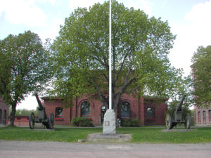 Punatiilinen vanha kasarmirakennus, jonka edessä lipputangon edessä oleva muistokivi.
