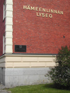 Punainen tiilirakennus, jonka seinässä lukee Hämeenlinnan lyseo. Nimen alla muistolaatta.