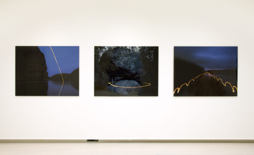 Ripustuskuva Pekka Luukkolan kolmesta valokuvasta. Teoksissa on kuvattu tulta pitkällä valotusajalla luonnon keskellä.