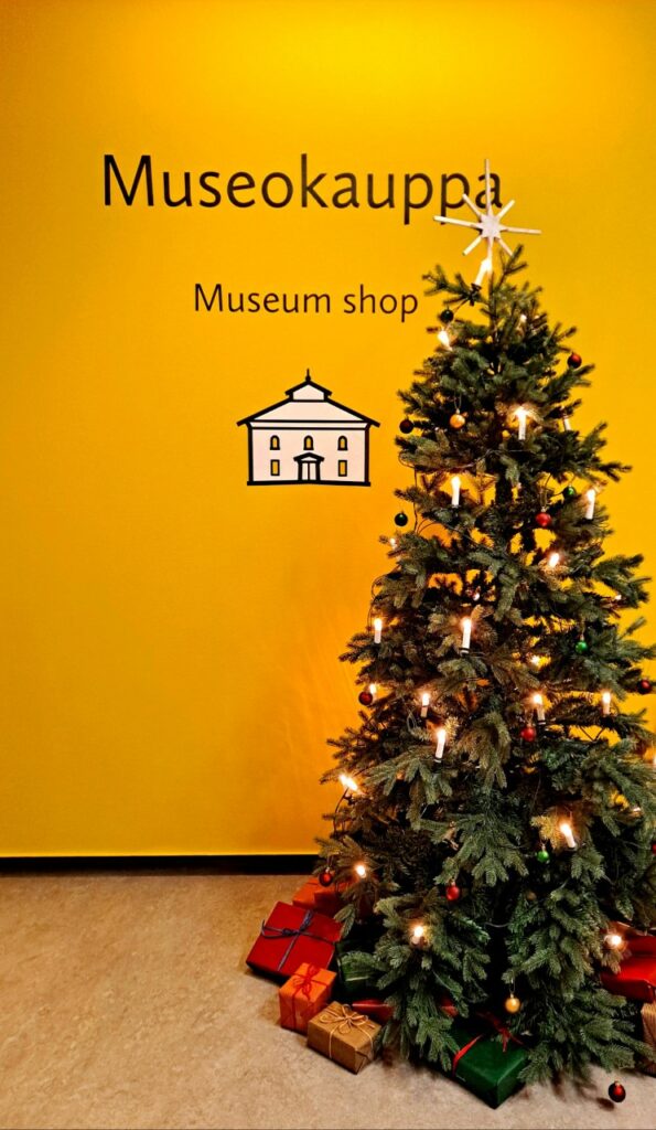 Joulukuusi keltaisen seinän edessä. Seinässä teksti museokauppa. Kuusi koristeltu kynttilöin ja kuusen juurella paketteja.